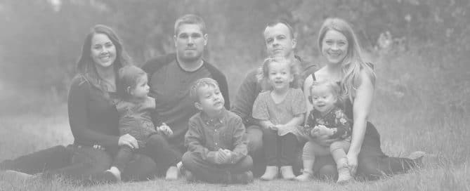 A group of two men, two women, and four young children sit together outdoors on a grassy path, posing for a photo—just the kind of happy moment you want to protect. For peace of mind at home, contact Family Plumbing.
